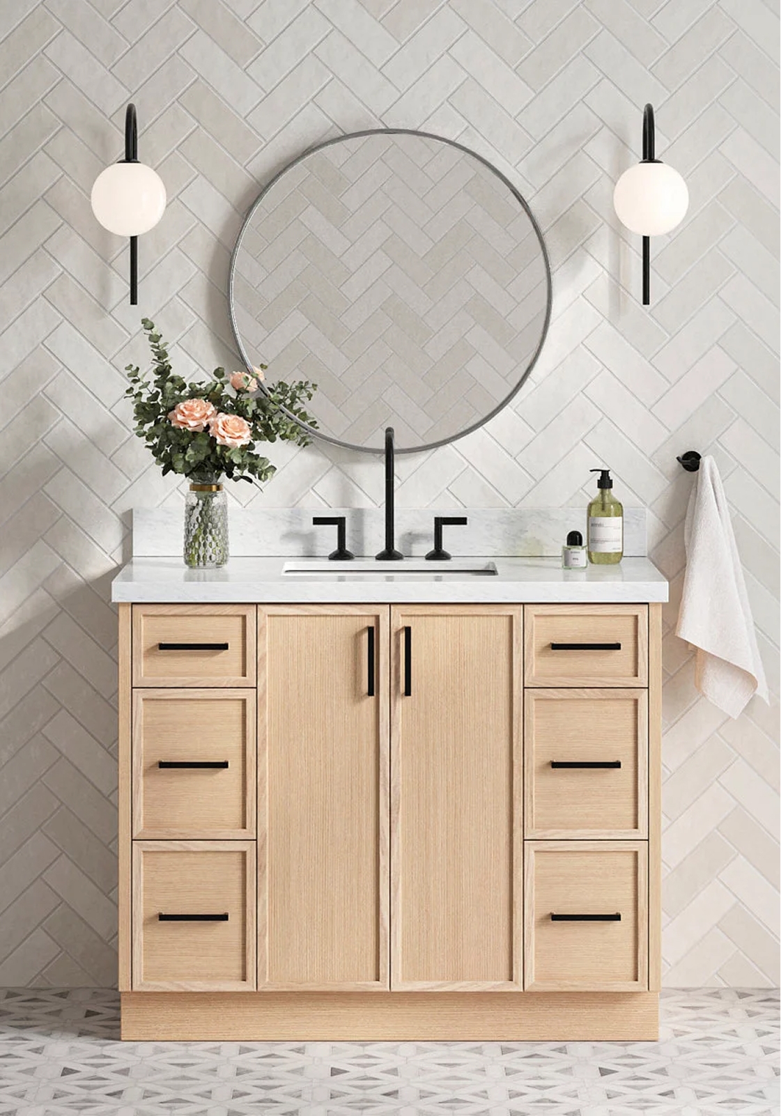 White oak bathroom vanity Light oak vanity with black hardware, round mirror, marble top, and floral decor styled against herringbone tile.