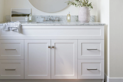 White double vanity with marble top and chrome hardware, styled with round mirror, potted plant, and towel on side.